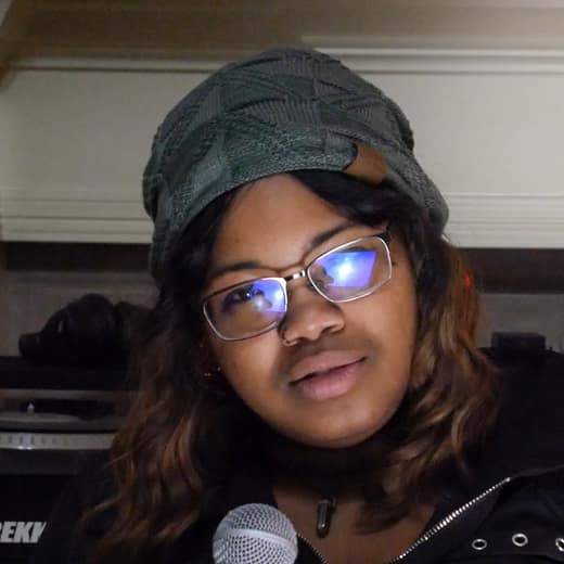 Tiffany Huff in a green beanie and glasses at the mic, turntables behind her
